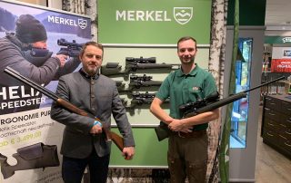 Ein Jagsschule Lüdersburg Mitarbeiter steht mit einem Jäger an einem Merkel Gewehre Stand in einem Fachgeschäft.